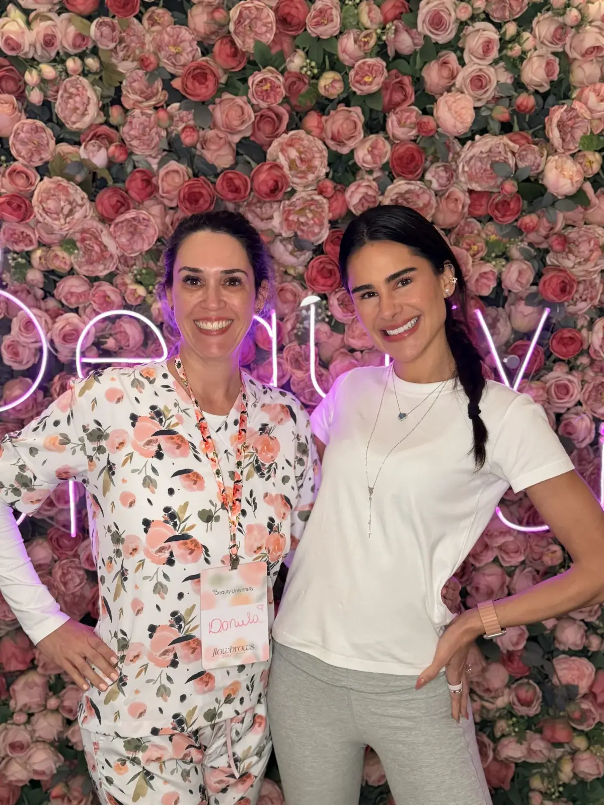 Daniela Rodrigues e a instrutora Natalia Martins posando em frente ao painel de flores da Beauty University durante o curso Flow Brows